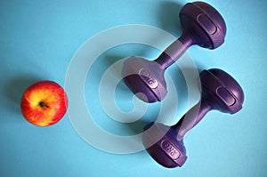 Dumbbells near red apple on blue background. Healthy lifestyle and sports concept. Apple fruit and blue barbell. Health regime and
