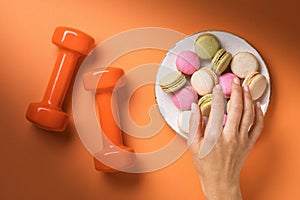 Dumbbells on the table and the hand of a man stretches behind macaroons in a plate, a top view. Concept on healthy lifestyle