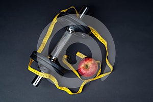 Dumbbells, tape measure and an apple on a dark background
