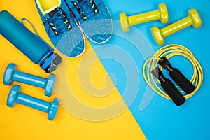 Dumbbells, trainers, jumping rope and a water bottle on blue and yellow background