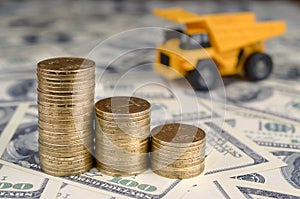 Dump truck toy and stacks of gold coins on background of many hundred dollar bills