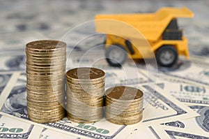 Dump truck toy and stacks of gold coins on background of many hundred dollar bills