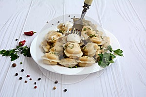 Dumplings on a white plate with fork, greens and sour cream on a white wooden table