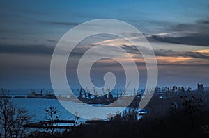 Dusk settles over harbor with silhouette of cranes against twilight sky. Industrial seascape at sunset, city skyline