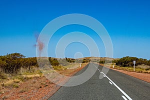 Dust devil besides dark road leading through red sanded Australian landscape towards Karijini National Park