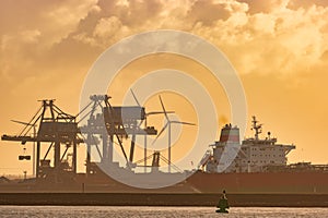 Dutch industrial plant with chimneys and windmill with cargo ship in front during sunset in Europoort, Rotterdam harbor