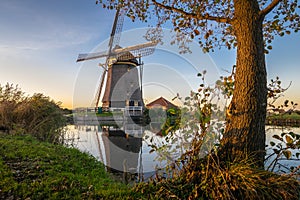 Dutch windmill along a canal with tree at sunset.