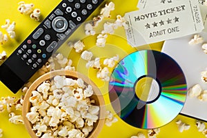 DVD or blu ray movie disc with tv remote control, movie tickets and bowl of popcorn on yellow background. Home theatre movie or