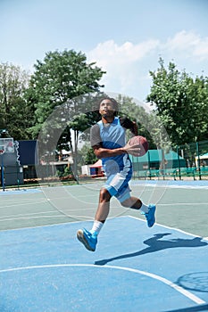 Dynamic image of young man, basketball player in blue uniform, training outdoors on playground on warm sunny day