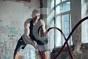 Dynamic image of young sportive man with strong muscular fit body training, doing exercises with battle ropes indoors