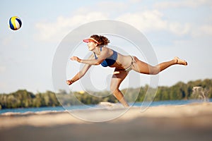 Dynamic image of young woman in motion, playing beach volleyball, hitting ball and falling down on sand