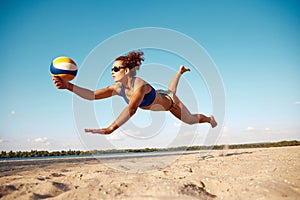 Dynamic image of young woman in motion, playing beach volleyball, hitting ball and falling down on sand. Sunny warm day