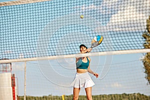 Dynamic image of young woman playing beach tennis, hitting ball with racket. Outdoor training on warm summer day