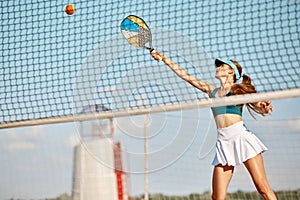 Dynamic image of young woman playing beach tennis, hitting ball with racket. Outdoor training on warm summer day