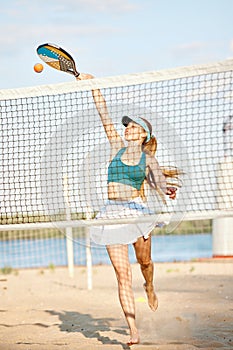 Dynamic image of young woman playing tennis, hitting ball with racket in motions. Beach training on warm summer day