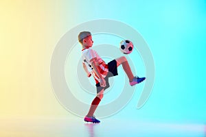 Dynamic photo of little athletic boy, soccer player kicking ball with knee in motion in neon light against gradient