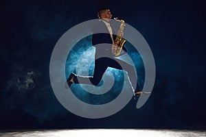 Dynamic shot of artistic man, solo performer jumping while playing jazz melody on saxophone against dark background with