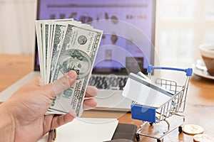 E-commerce concept, hand holding money and credit card in shopping cart with laptop open shopping website background