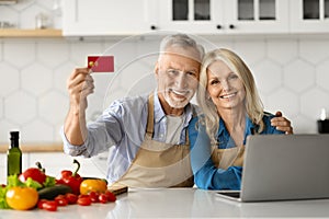 E-Commerce Concept. Senior Couple Holding Credit Card And Using Laptop In Kitchen