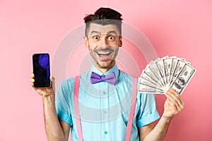 E-commerce and shopping concept. Cheerful guy in bow-tie showing empty smartphone screen and dollars money, smiling