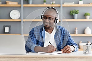 E-education. Happy black mature man using laptop and taking notes, listening online course