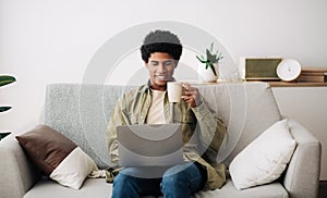 E-learning concept. Handsome black male student using laptop computer and drinking hot coffee at home, panorama