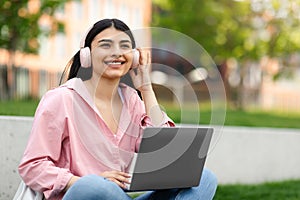 E-learning concept. Happy hispanic teen lady using laptop, wearing headphones listening to music, sitting in campus park