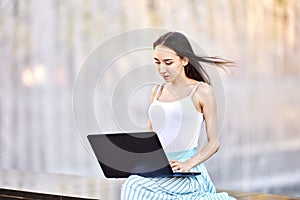 E-Learning or Telecommuting using laptop connected to Internet, young woman does remote work while visiting public park.