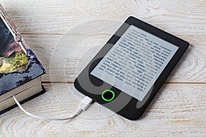 E-reader connected by cable to a paper book on a white wooden table. Copy space on e-book display. E-reading on anti-glare screen photo