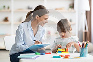 Early kids development. Cute little boy playing logical geometry game, woman specialist with notepad watching him