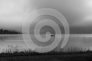 An early morning at Lac de Joux, Switzerland: A lone fisherman on a boat, fog is still hiding the rising sun