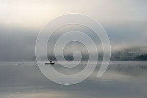 An early morning at Lac de Joux, Switzerland: A lone fisherman on a boat, fog is still hiding the rising sun