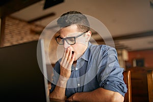 Early morning in the office. Sleepy tired handsome hipster freelancer in glasses is yawning at his work place in front