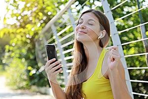 Early morning portrait of a happy young fitness girl sitting in the park enjoying listening to music using mobile phone with