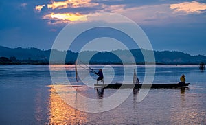 In the early morning before sunrise, an Asian fisherman on a wooden boat casts a net for catching freshwater fish in a natural