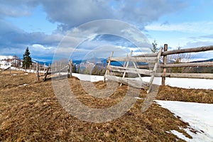 Wooden gate on snowcovered pasture, ark clouds over mountains.