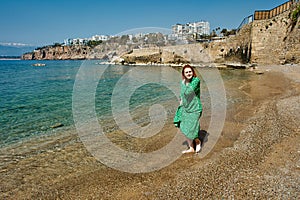 Early spring in turkish antalya, young white woman in green evening dress walks barefoot along beach.