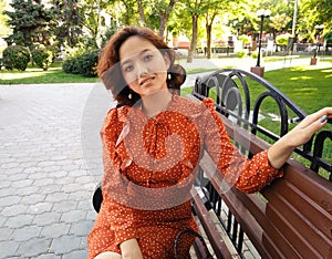 East Asian woman sitting on park bench smiling happy at camera. Pretty young japanese woman enjoying summer in park on parkbench