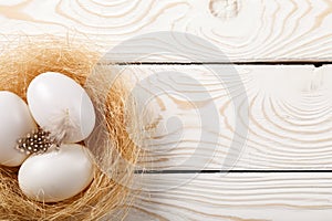 Easter background. Easter white eggs and feather in nest on rustic white wooden background. Top view, copy space