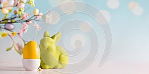 Easter banner with a yellow egg and a green rabbit on a blue background with a bokeh and a branch of flowers. Copy space