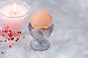 Easter brown egg in a crystal blueish cup, burning pink candle, tender red spring flowers