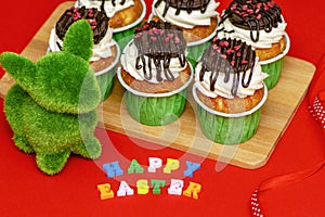 Easter bunny made of grass and delicious bright cupcakes on a wooden board on a red background.