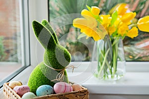 Easter bunny rabbit statuette in straw basket with colored eggs on the windowsill with fresh spring tulips and daffodils