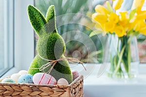 Easter bunny rabbit statuette in straw basket with colored eggs on the windowsill with fresh spring tulips and daffodils