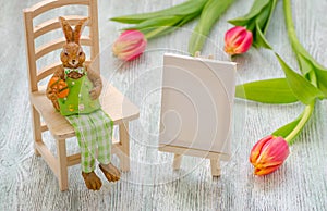 Easter bunny sitting on the stool with a egg, painting easel and tulips over wooden background