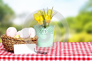 Easter card template. Colorful easter eggs in a wicker basket, yellow tulips and a empty tag on a red checkered tablecloth against