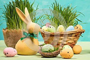 Easter card. Yellow bunny, colorful eggs on table, in nest, in basket with green grass on turquoise.