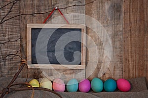 Easter colored eggs with bow and black board with copy space for text against natural wooden textured background on linen fabric.