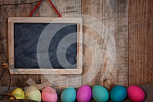 Easter colored eggs with bow and black board with copy space for text against natural wooden textured background on linen fabric.