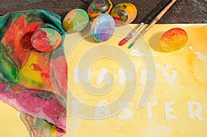 Easter colorful eggs with two painter brushes and a hand painted cloth,arranged on watercolor paper with yellow painted text.